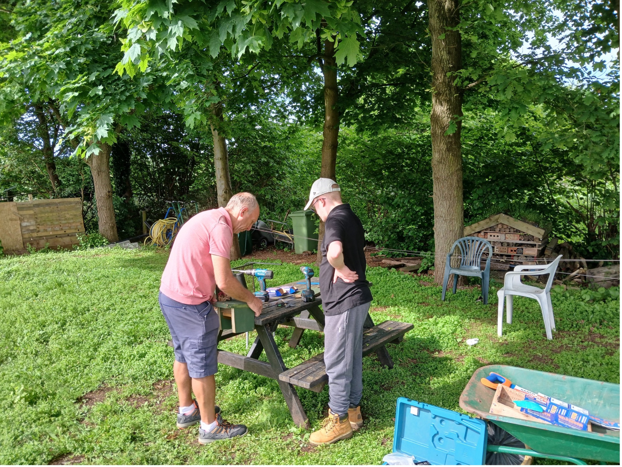 <br />
Two men are working outdoors on a picnic table in a lush, green garden. One man, in a pink shirt and shorts, is bent over the table, concentrating on a project, while the other man, in a black shirt and grey pants, stands nearby observing. The table is covered with various tools and equipment. The garden is surrounded by tall trees providing shade, and there are plastic chairs and a wheelbarrow in the background.