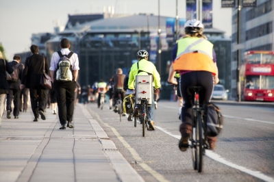 Cyclists and pedestrians commuting on a busy city street during peak hours, with a red double-decker bus and cars in the background.