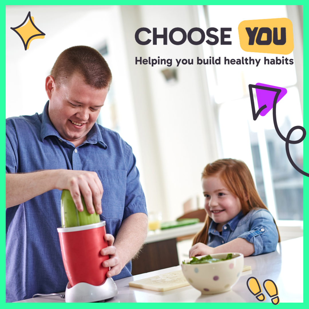 A man and a child, at a kitchen counter. The man is using a blender to blend a green liquid. There's a bowl on the counter in front of him containing green leafy veg.