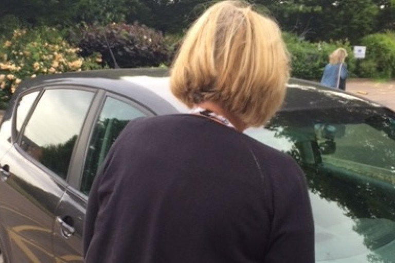 Person standing beside a parked car in a roadside parking bay, viewed from behind during an inspection.
