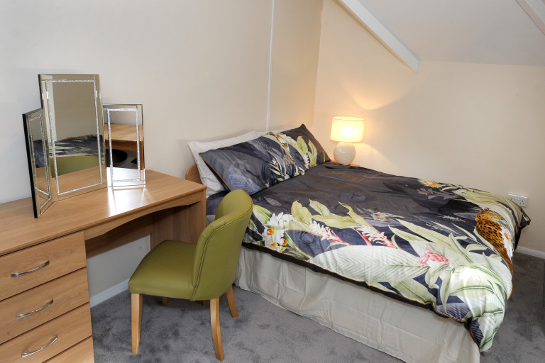 Furnished bedroom with double bed, desk, chair, bedside lamp and soft grey carpet.