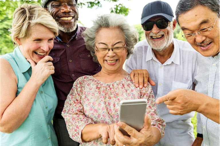 Group of five adults gathered outdoors, looking at and sharing a smartphone screen together.