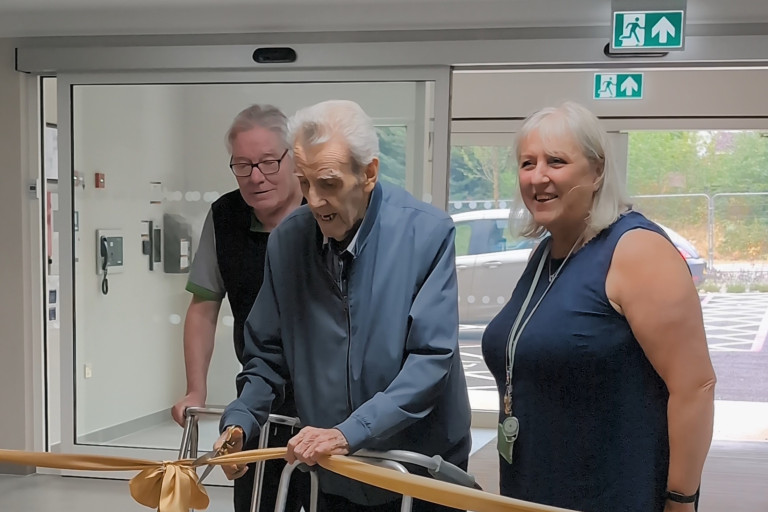 Person using a walker cuts a gold ribbon at Honeysuckle House opening, flanked by two attendees near entrance doors.