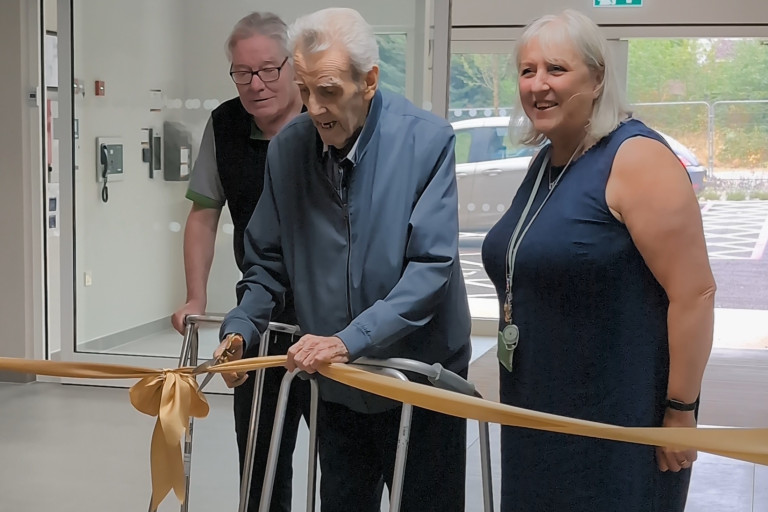 Person using a walker cuts a gold ribbon at Honeysuckle House opening, flanked by two attendees near entrance doors.