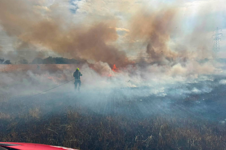 Firefighter battling a grass fire in a smoke-filled field, with flames and power lines visible.