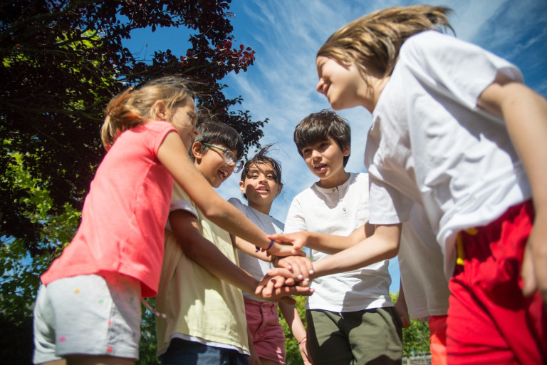 Five children outdoors in a circle, stacking hands together during a team activity under a sunny blue sky.