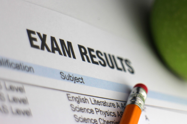 Exam results sheet showing subject grades, with a pencil pointing at the list and a blurred green apple nearby.