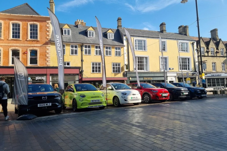 Line of display cars in a town square outside historic shopfronts under a clear blue sky.