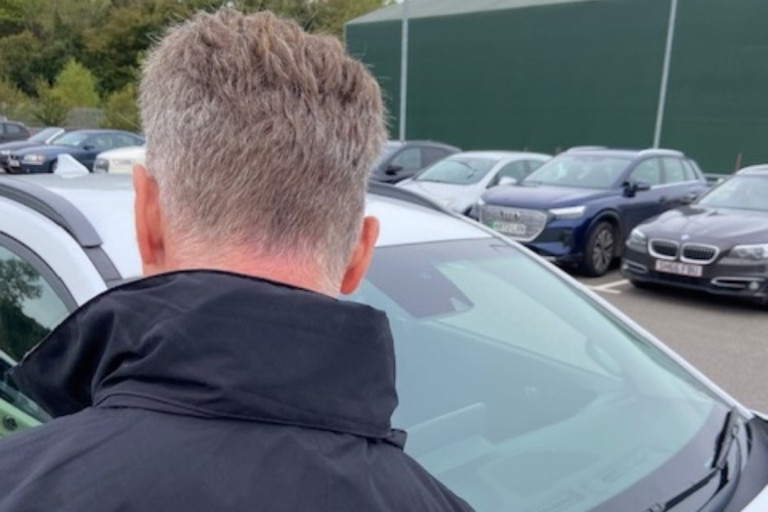 Person viewed from behind leaning towards a parked car in a car park, with several vehicles in the background.