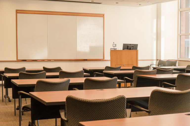 Empty classroom with rows of desks and chairs facing a whiteboard and lectern near large windows.