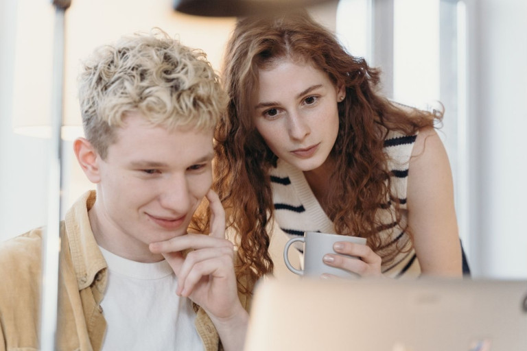 Two people looking at a laptop together indoors, one holding a mug while reviewing content on screen.