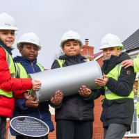 Children in hard hats and hi-vis vests hold a time capsule at a construction site
