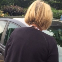Person standing beside a parked car in a roadside parking bay, viewed from behind during an inspection.