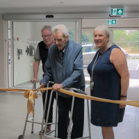 Person using a walker cuts a gold ribbon at Honeysuckle House opening, flanked by two attendees near entrance doors.