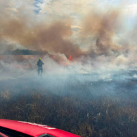 Firefighter battling a grass fire in a smoke-filled field, with flames and power lines visible.