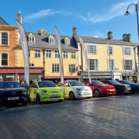 Line of display cars in a town square outside historic shopfronts under a clear blue sky.