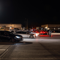 Cars gathering on an industrial estate at night, with a cyclist riding past traffic cones.