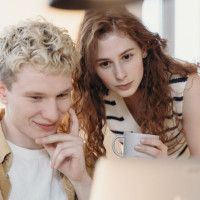 Two people looking at a laptop together indoors, one holding a mug while reviewing content on screen.