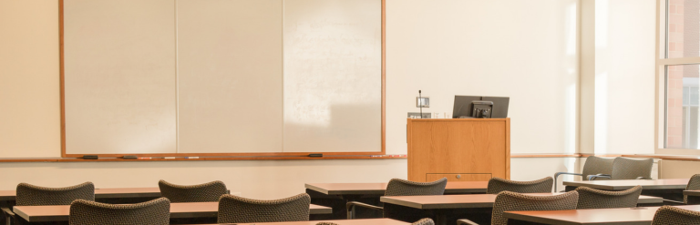 Empty classroom with rows of desks and chairs facing a whiteboard and lectern near large windows.